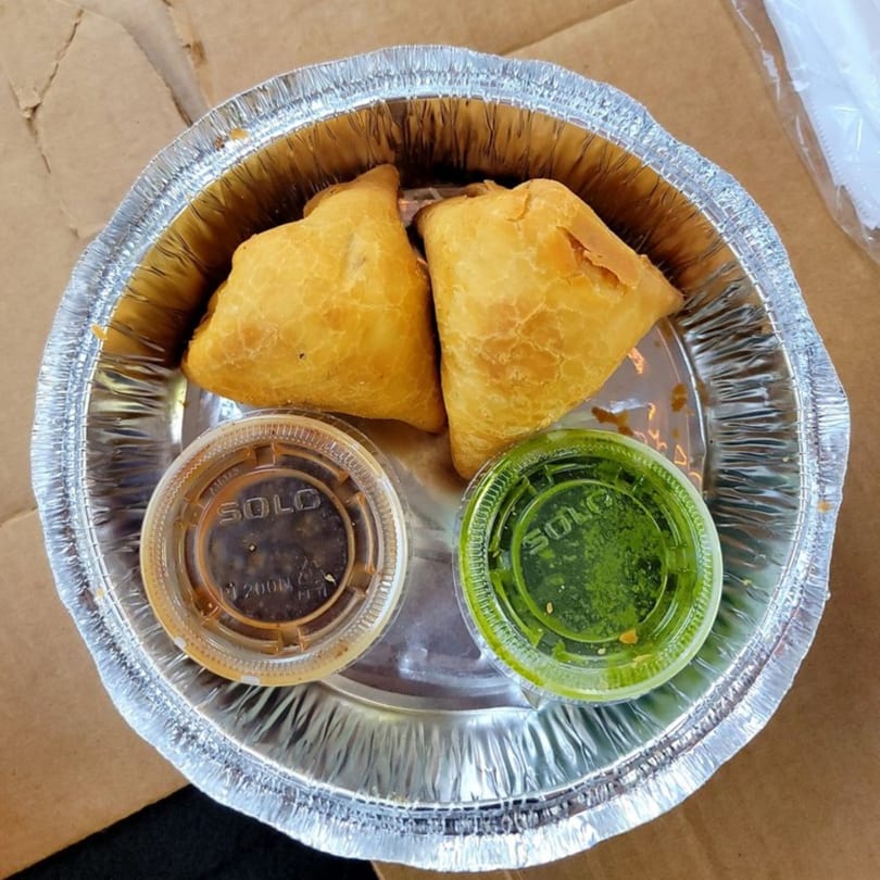 Two golden-brown Indian samosas with brown and green dipping sauces in a foil takeout container.