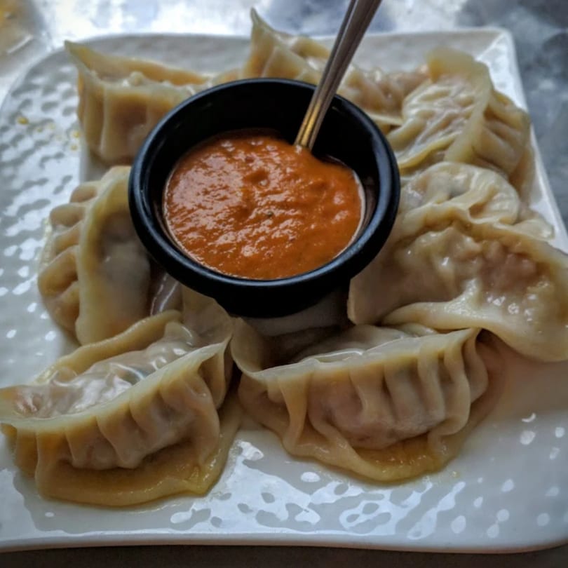 A plate of Indian momos, steamed dumplings, with a small black bowl of red dipping sauce.